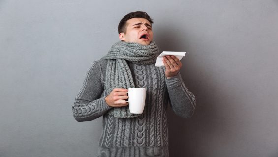Sick Man in sweater and scarf sneezes while holding tea